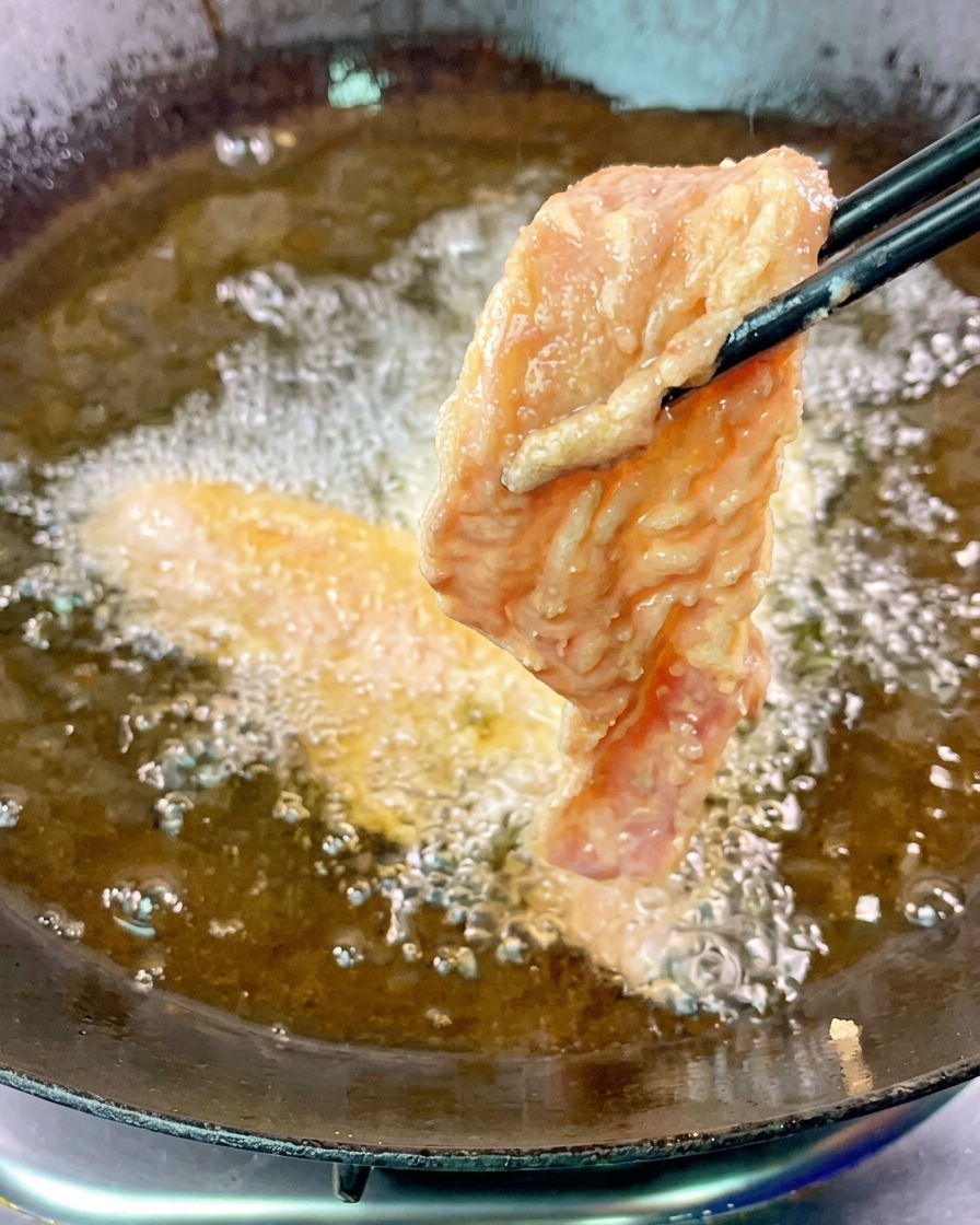 A single slice of battered pork belly being lowered into shimmering hot oil with black chopsticks.