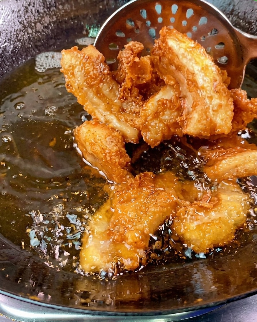 Crispy golden brown pork belly slices being lifted out of a bubbling wok of oil with a slotted metal spoon.
