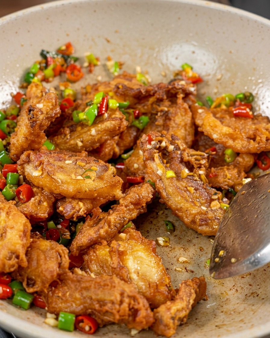 A close-up of crispy pork belly, red and green peppers, and garlic being tossed in a wok with white sesame seeds visible on the surface.