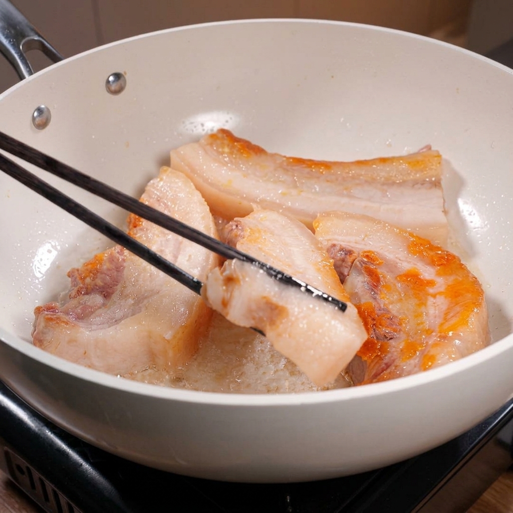 Long black chopsticks flip thick strips of pork belly in a white frying pan, showing beautifully seared, golden-brown edges.