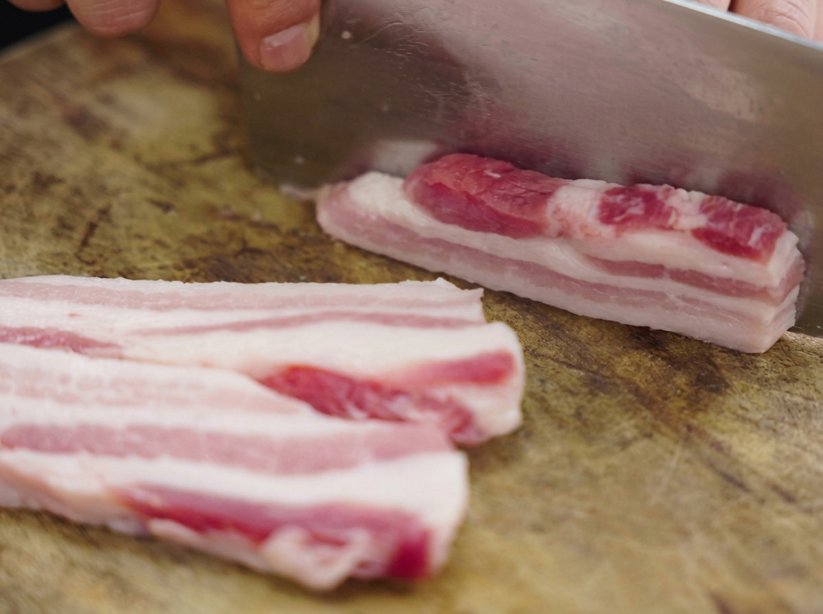 Thick strips of raw layered pork belly being sliced on a wooden cutting board with a large metal cleaver.