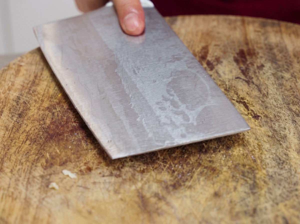 A heavy metal cleaver being pressed down flat onto a wooden cutting board to smash raw garlic cloves.
