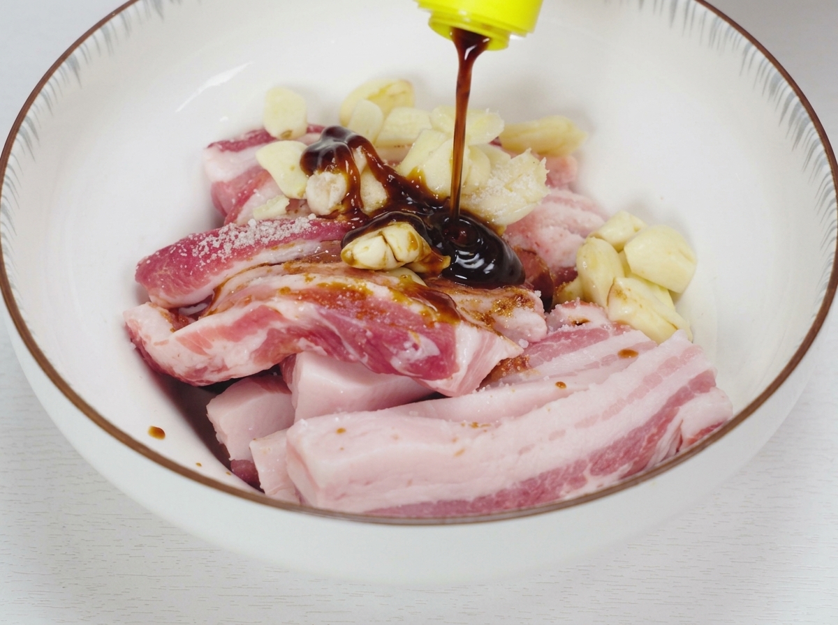 Dark liquid seasonings and oyster sauce being poured over raw sliced pork belly and smashed garlic in a white ceramic bowl.
