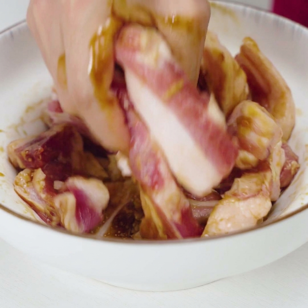 A hand vigorously mixing and kneading raw pork strips with a dark liquid marinade and garlic pieces in a white ceramic bowl.