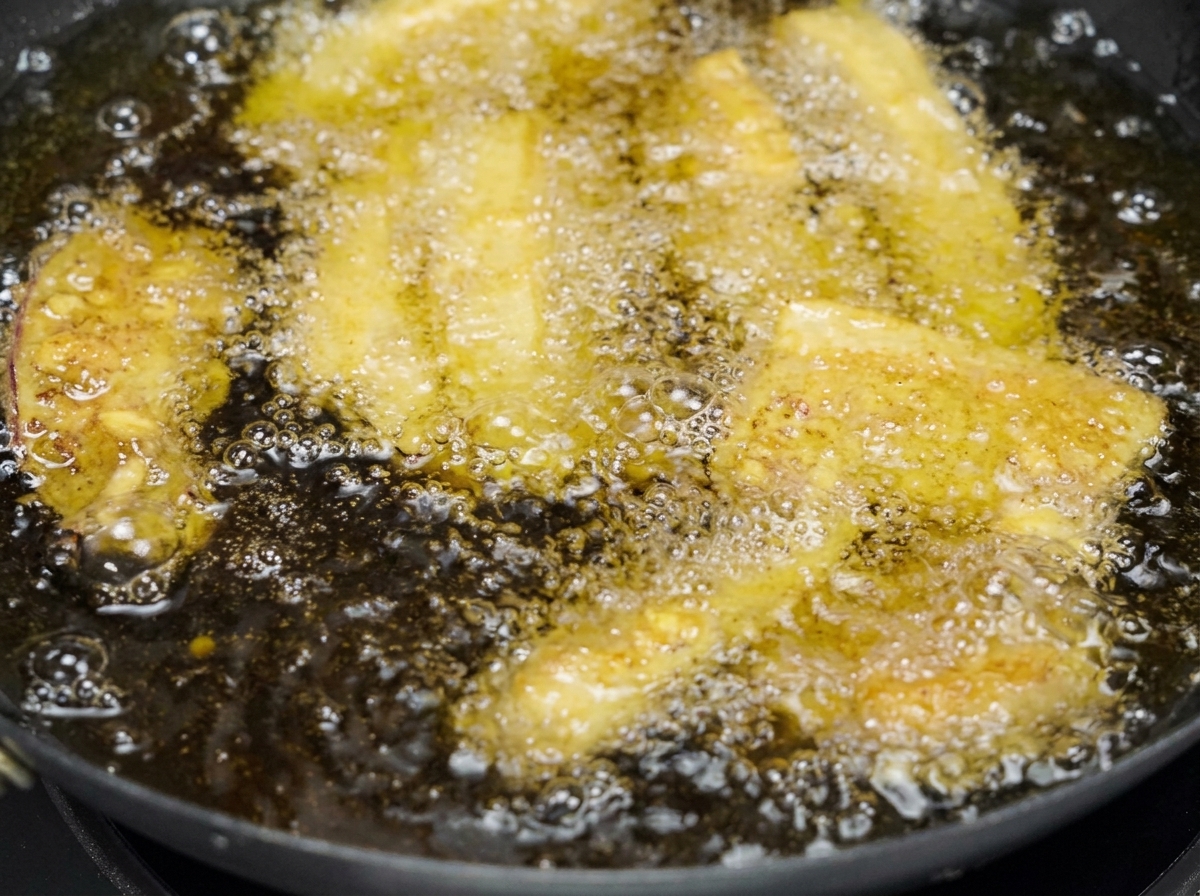 Several strips of coated pork belly deep-frying in a pan filled with bubbling golden cooking oil.