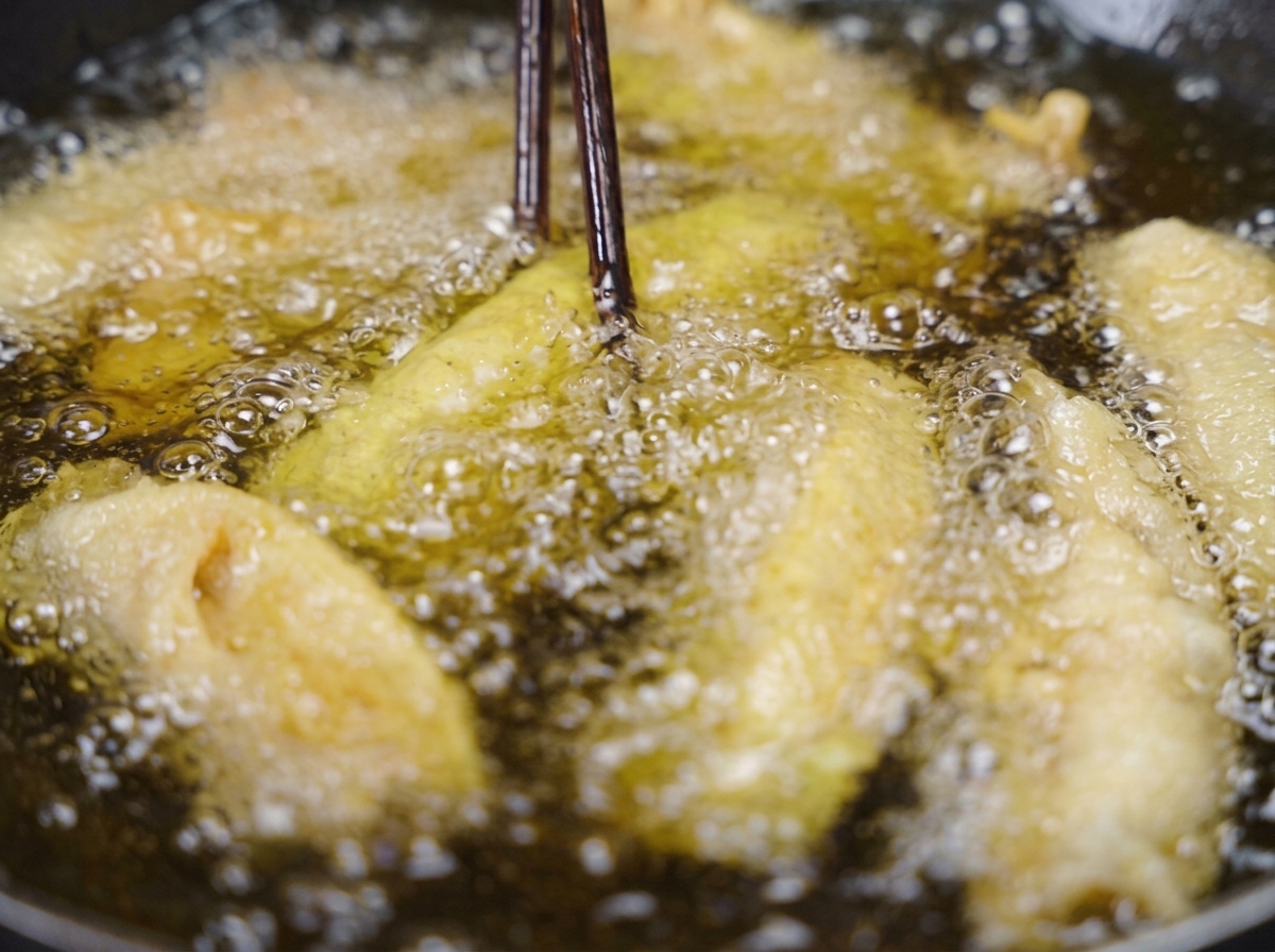 Dark wooden chopsticks lifting a crispy, golden-brown fried pork strip out of bubbling cooking oil.