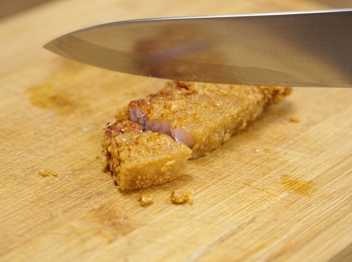 A large metal knife slicing a crispy, golden-brown piece of fried pork into smaller bite-sized sections on a wooden cutting board.