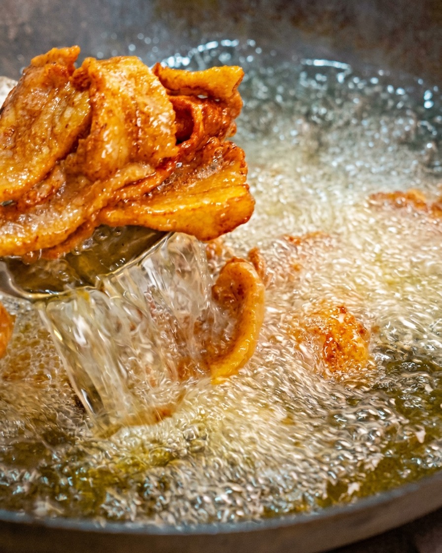Scooping golden-brown, curled slices of fried pork belly out of a wok of hot bubbling oil using a strainer ladle.