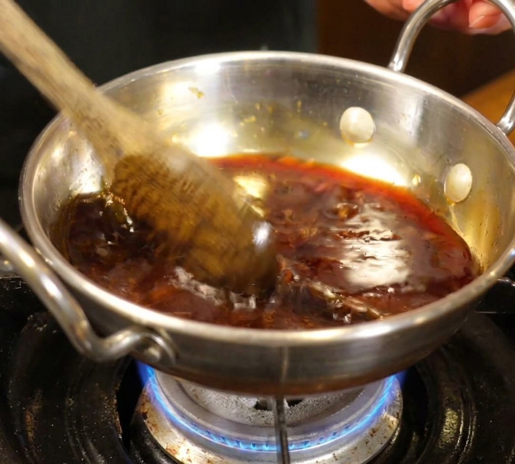 A wooden spoon stirring a dark, bubbling sauce mixture in a small stainless steel pot over a blue gas flame.