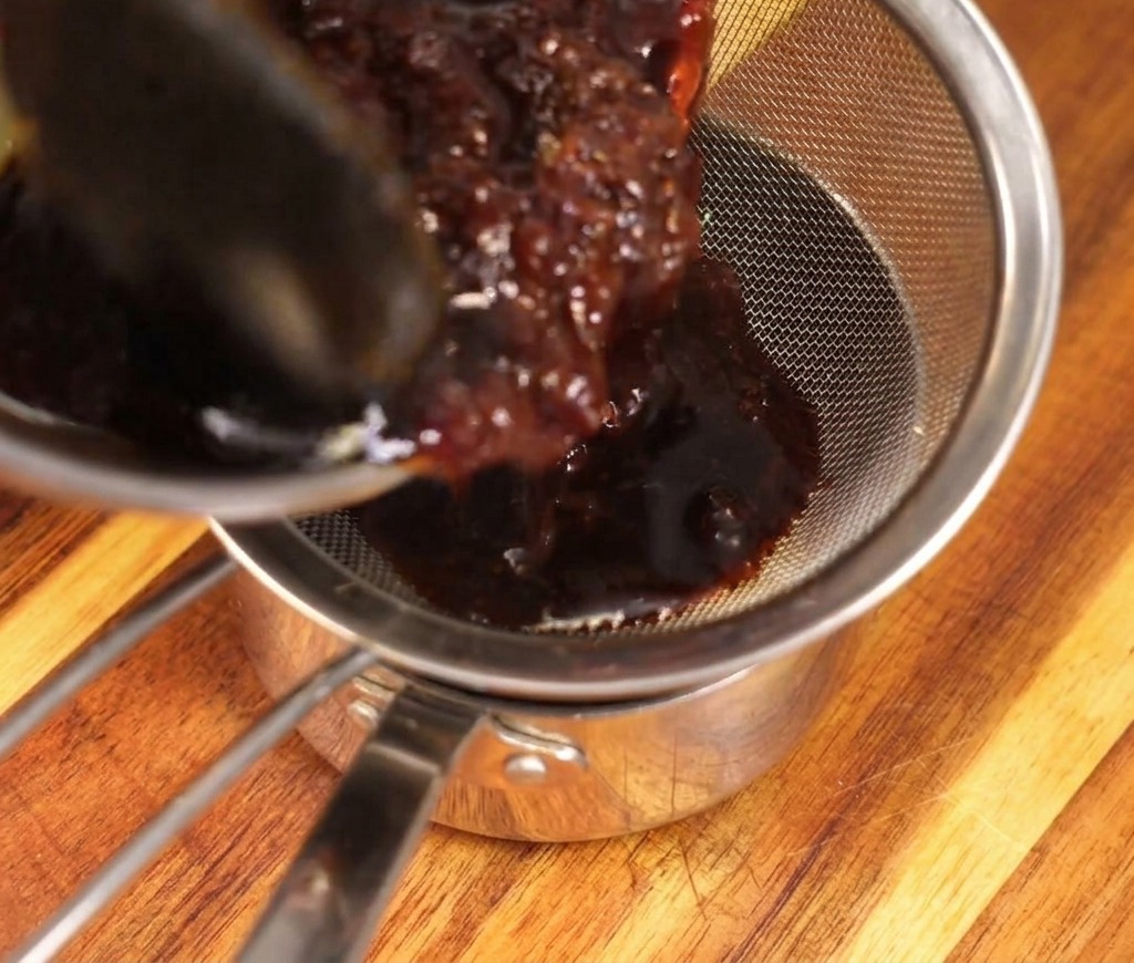 Rich, dark sauce being poured through a silver mesh strainer into a stainless steel pot, catching the soaked bonito flakes.