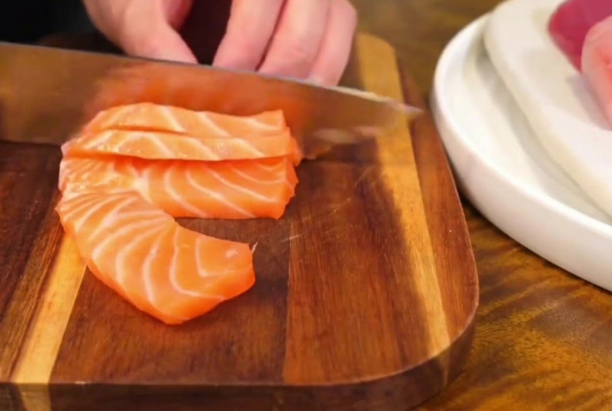 A sharp kitchen knife slicing through a fresh, marbled salmon fillet on a wooden board, creating thick sashimi slices.