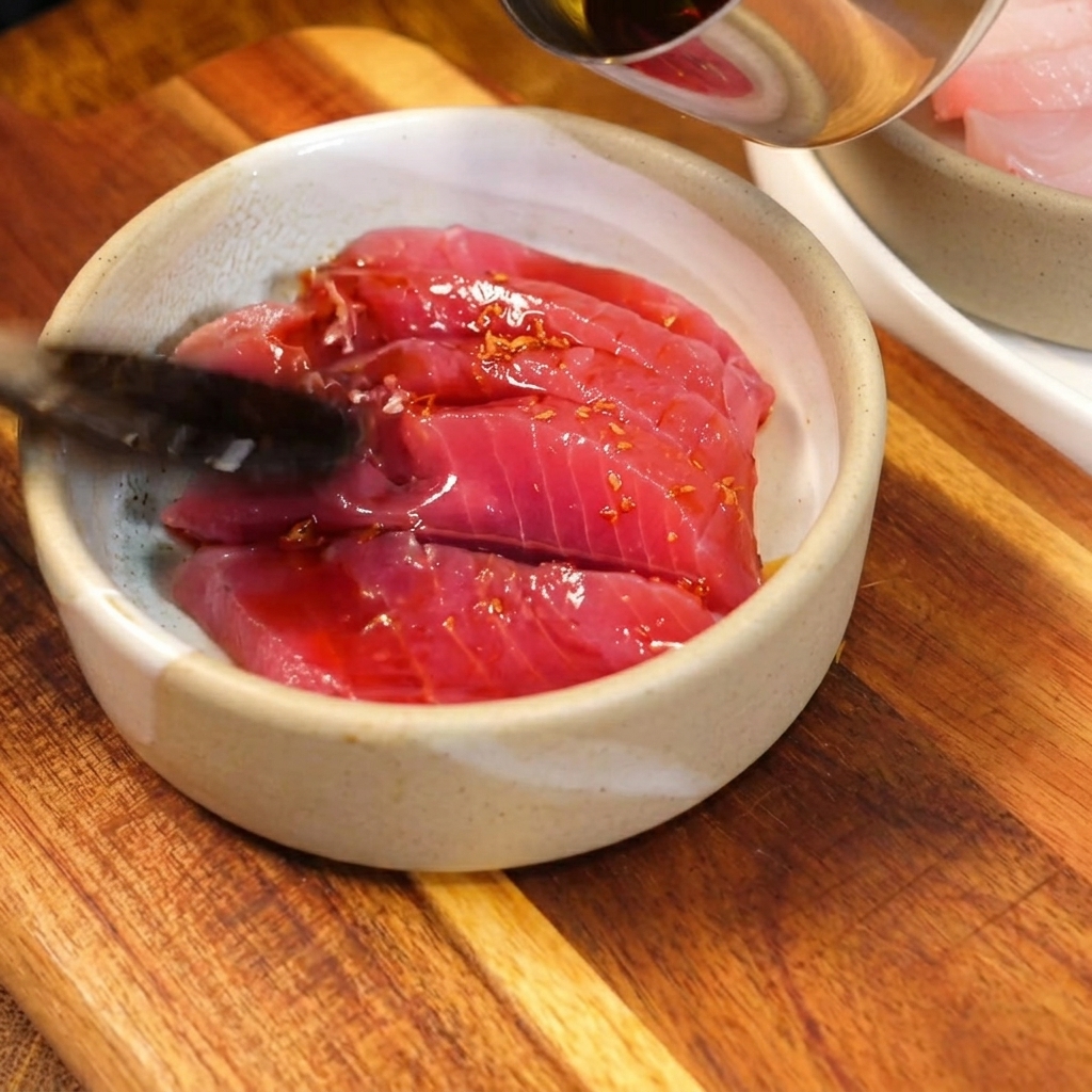 Dark, savory soy-based sauce being spooned over bright red, raw tuna slices in a small rustic ceramic bowl.