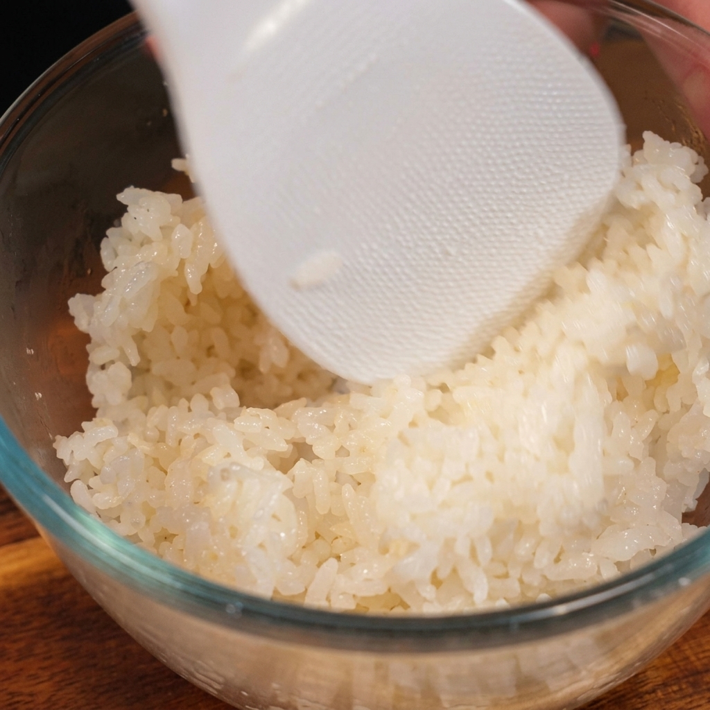 A white, textured plastic rice paddle gently folding seasoned, cooked white rice in a clear glass bowl.