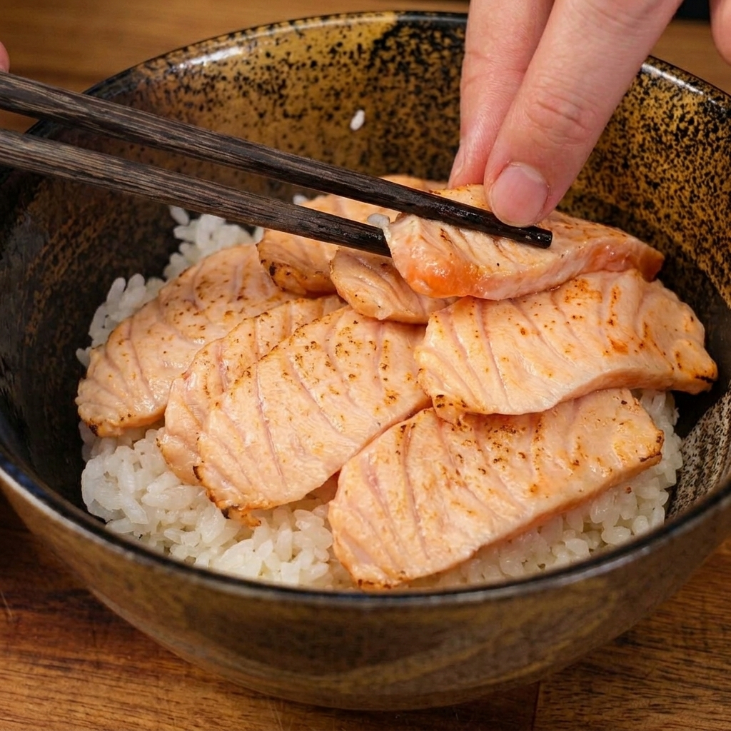 Wooden chopsticks gently placing a slice of lightly seared salmon onto a bed of white rice in a ceramic bowl.