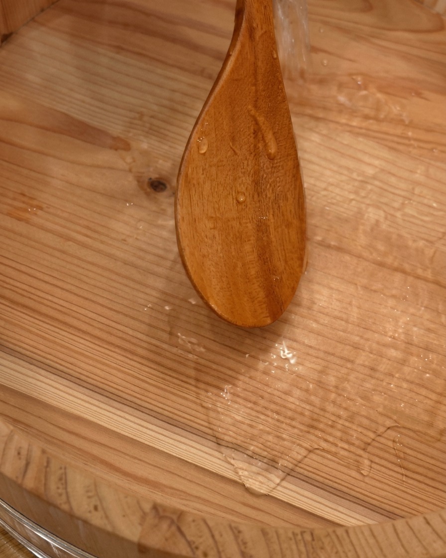 Water being poured from a wooden paddle into a traditional large wooden sushi mixing tub.