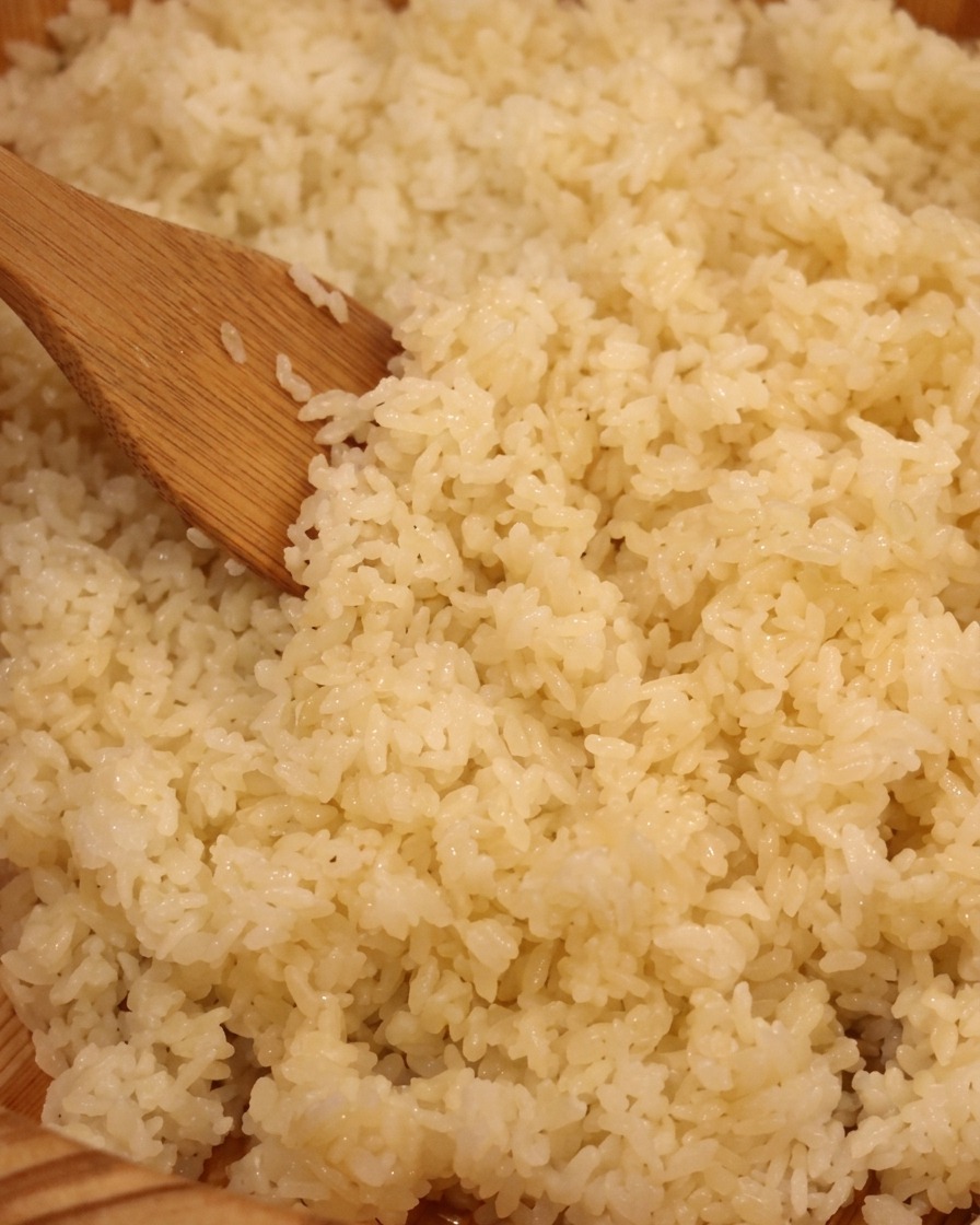 A close-up view of a wooden paddle mixing cooked white rice inside a traditional wooden sushi tub.