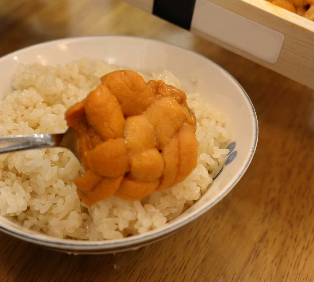 A spoon lifting a large, textured lobe of orange sea urchin to place it onto a bowl of white sushi rice.