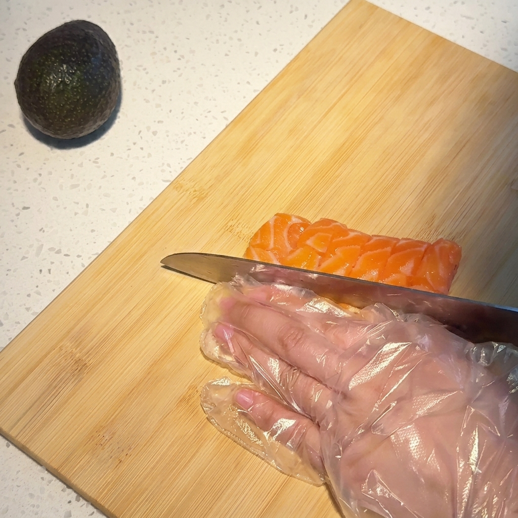 A close-up shot of a hand in a clear plastic glove using a chefs knife to slice a marbled salmon fillet into small, neat cubes on a bamboo cutting board.