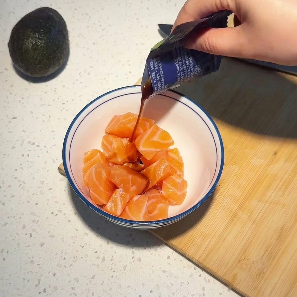 Dark soy sauce and wasabi seasoning being poured from a packet onto raw salmon cubes in a white bowl.