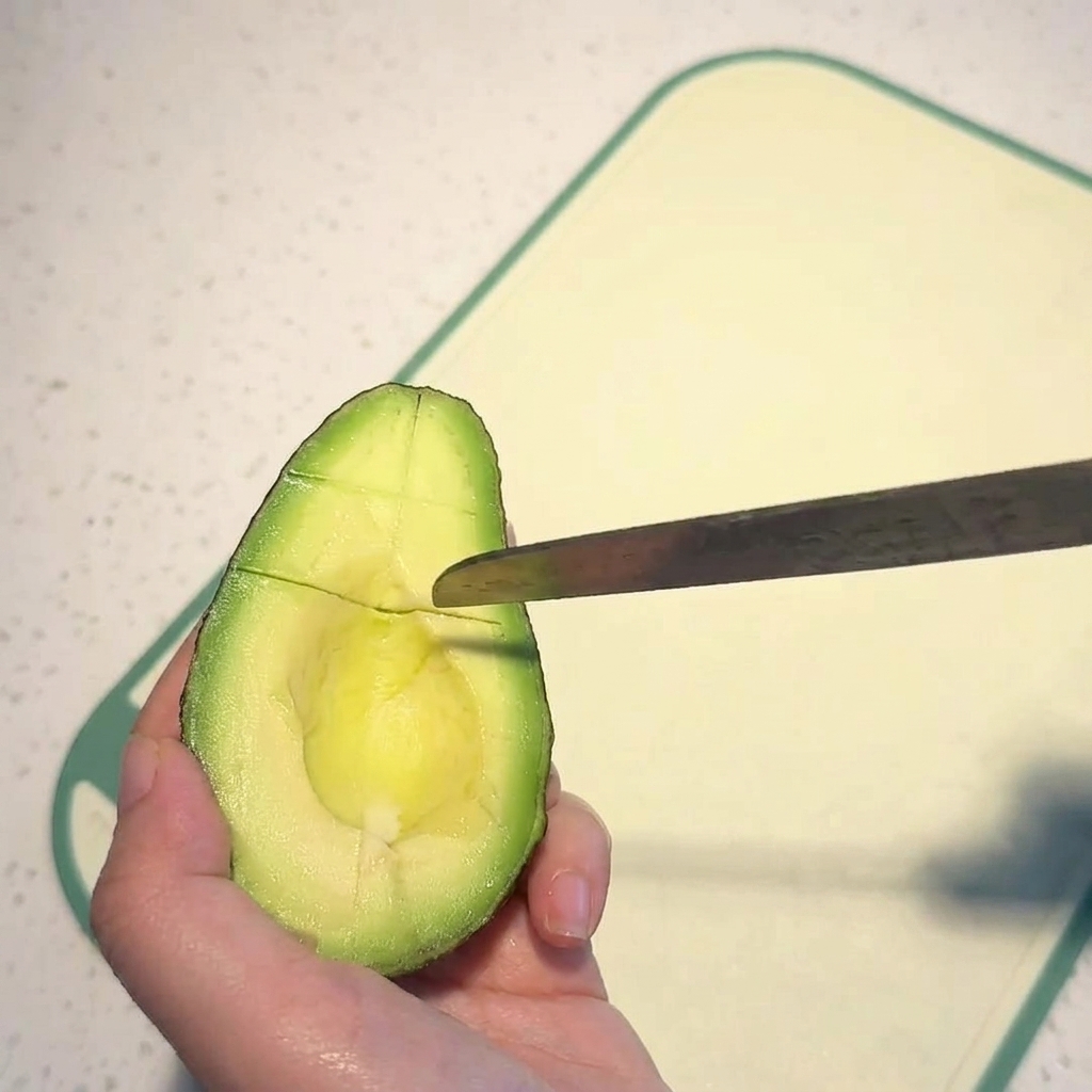 A hand holding an avocado half while a knife scores the bright green flesh into a grid pattern.