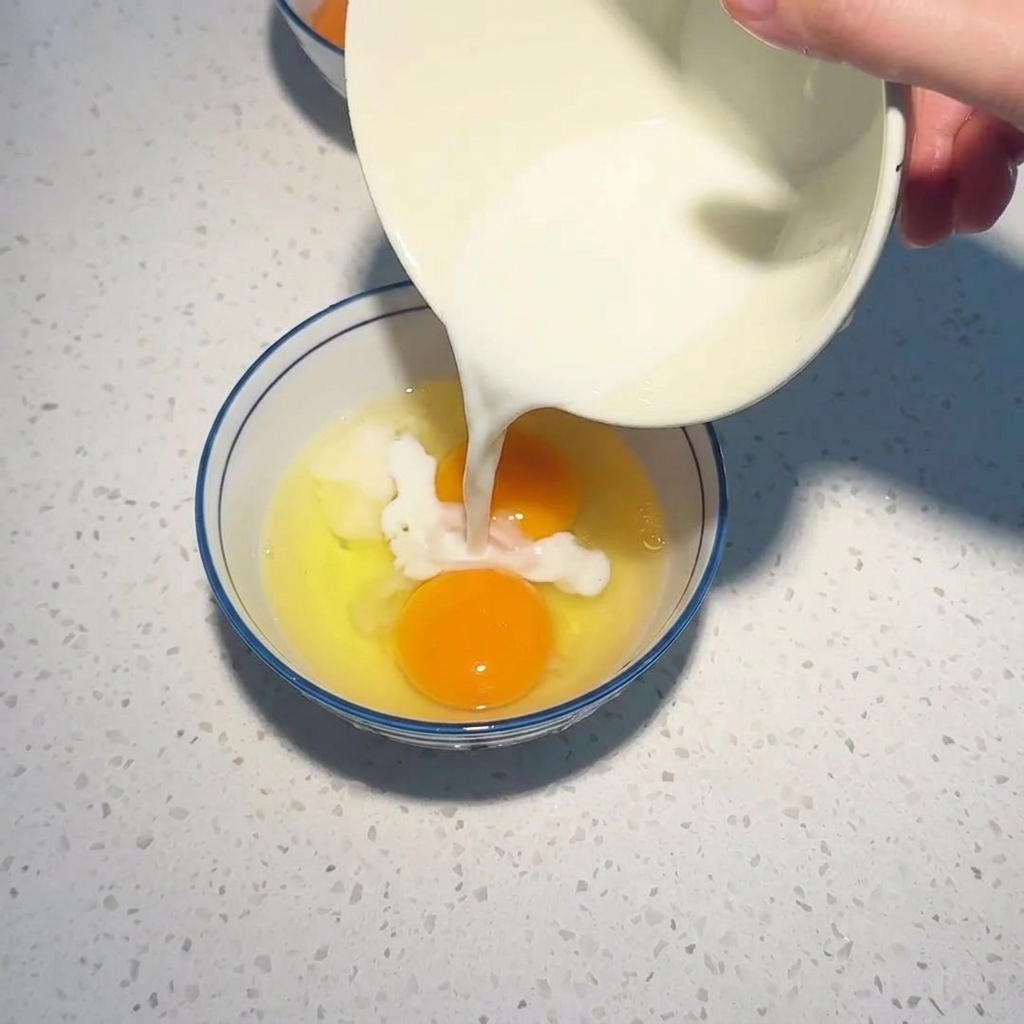 White liquid being poured from a bowl into another bowl with two cracked raw eggs.