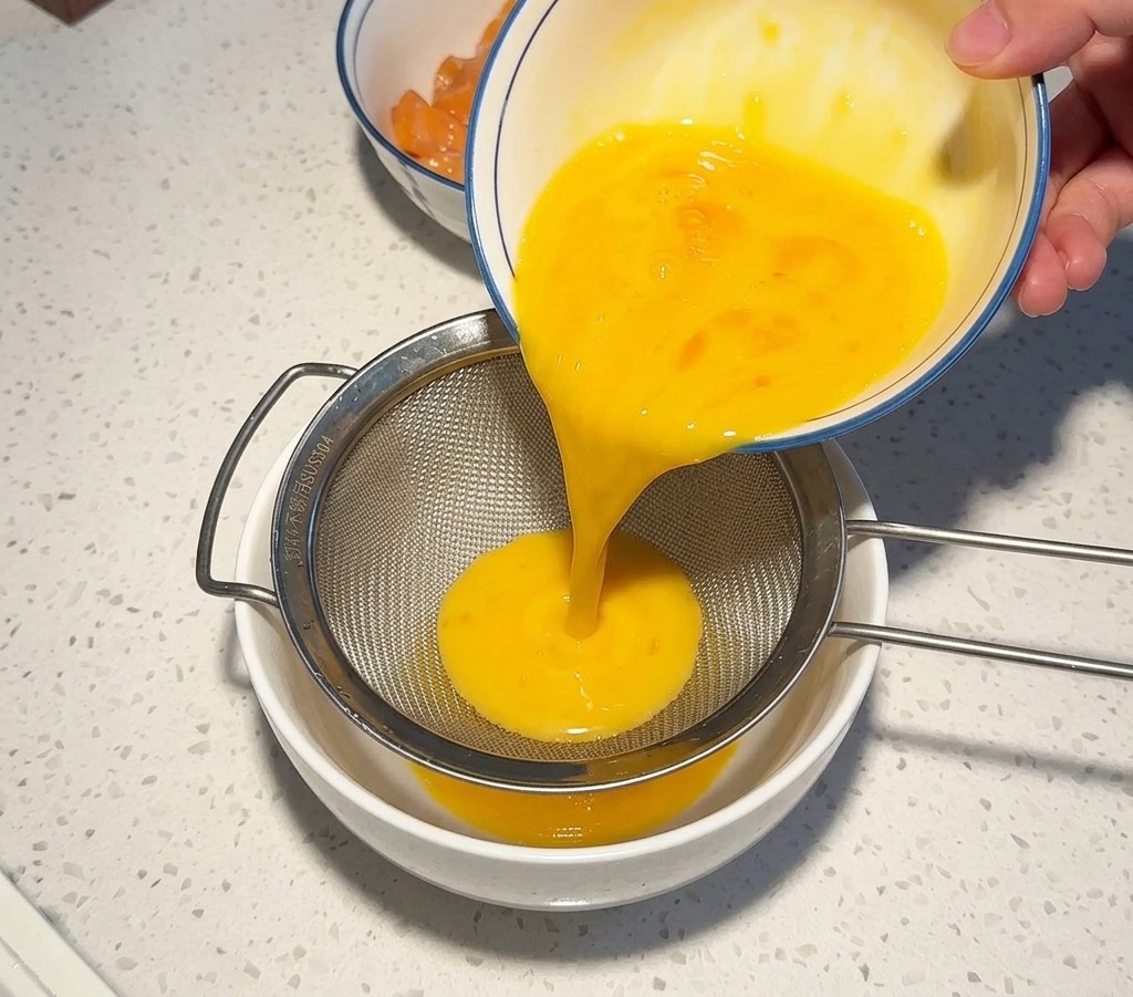 Beaten yellow egg liquid being poured from a bowl through a stainless steel sieve into a white bowl to ensure a smooth consistency.