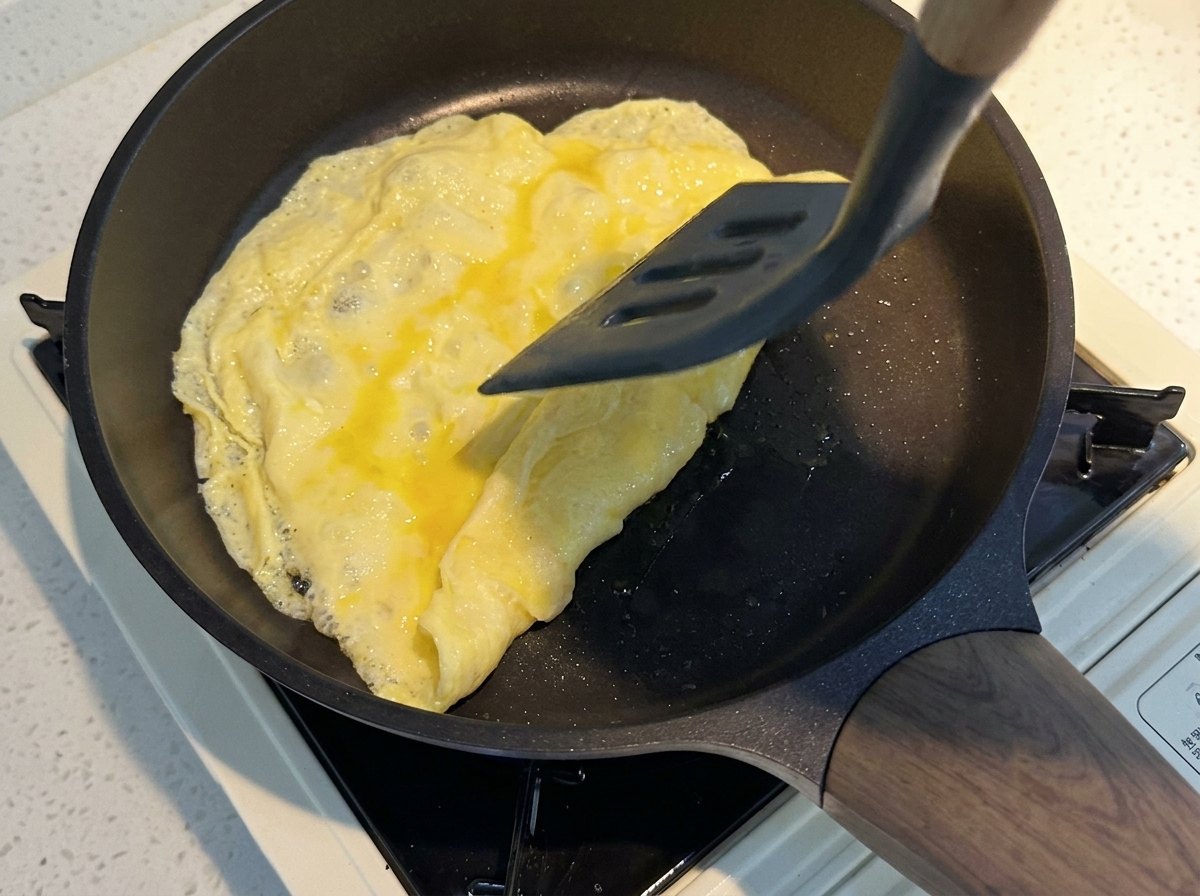 A grey spatula folding a thin layer of cooked yellow egg in a heated non-stick pan.
