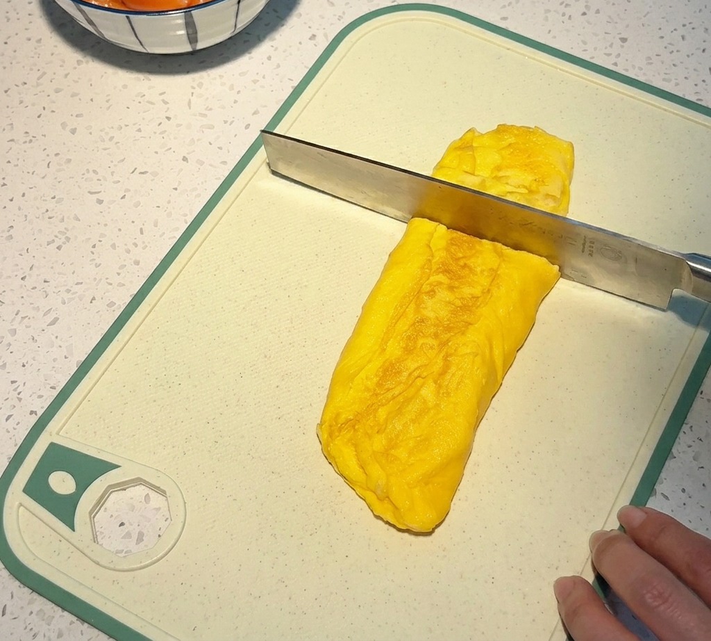 A knife slicing through a thick, golden-yellow rolled omelet on a white cutting board to create small square cubes.