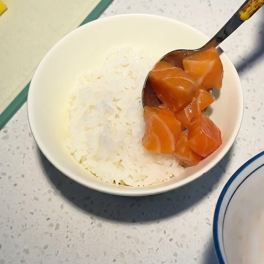 A metal spoon placing glistening, cubed raw salmon onto a bed of white rice in a bowl.