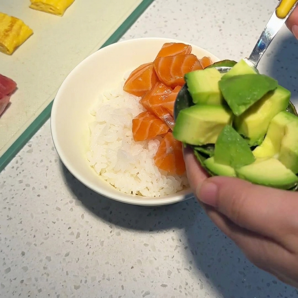 A hand using a spoon to scoop diced avocado cubes from their skin into a white bowl already containing rice and salmon chunks.