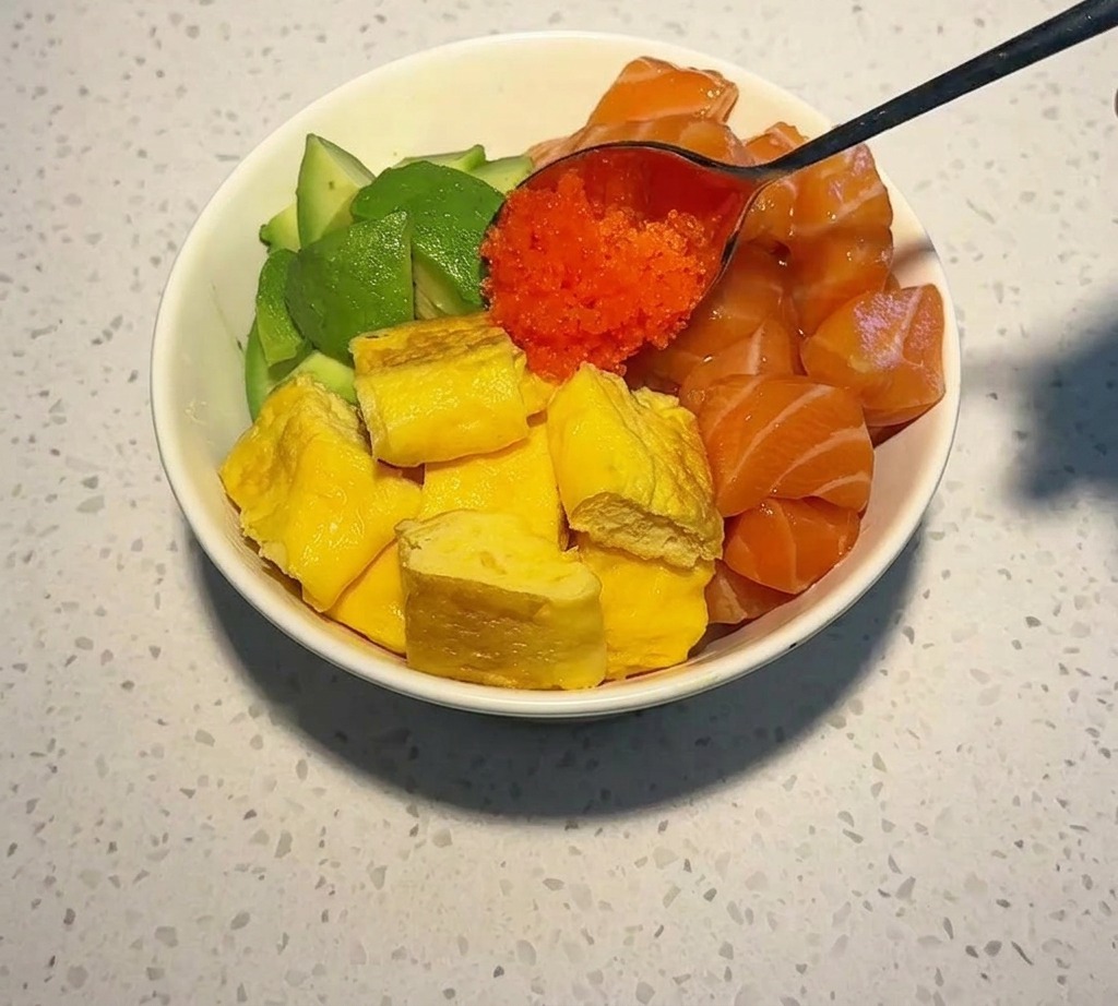 A metal spoon depositing a cluster of vibrant orange fish roe onto a bowl already filled with salmon cubes, avocado, and yellow egg.
