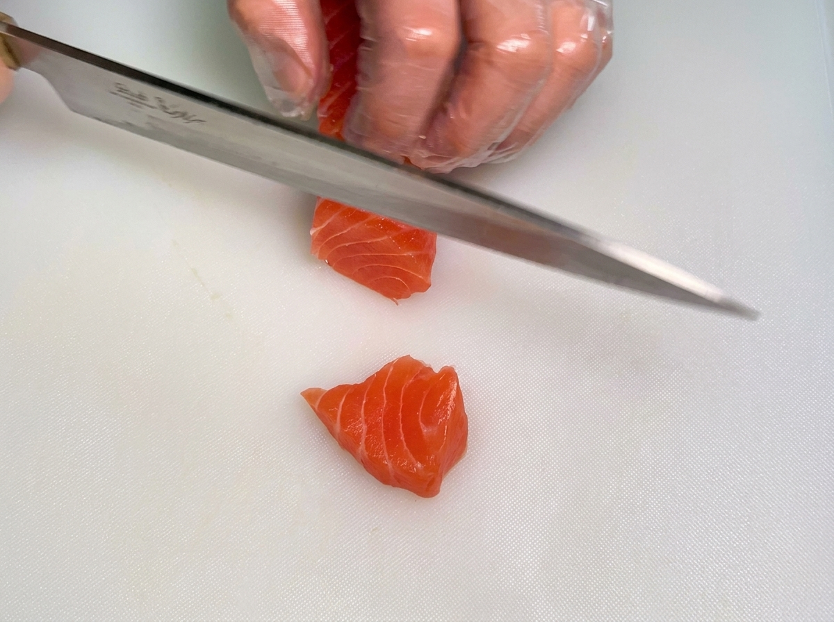 A chefs gloved hand holding a block of raw salmon while the other hand uses a knife to cut it into thick strips on a white board.