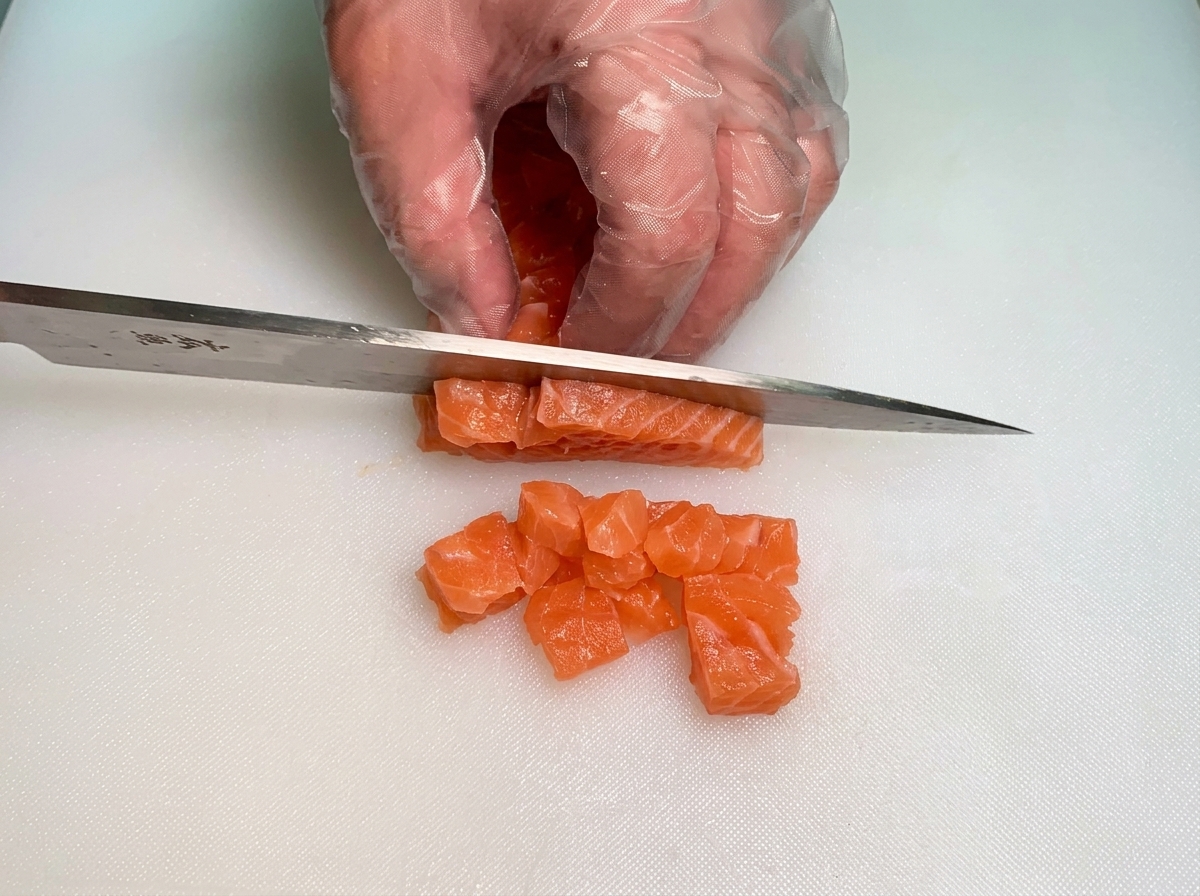 Gloved hands using a knife to dice thick strips of bright orange raw salmon into uniform small cubes.