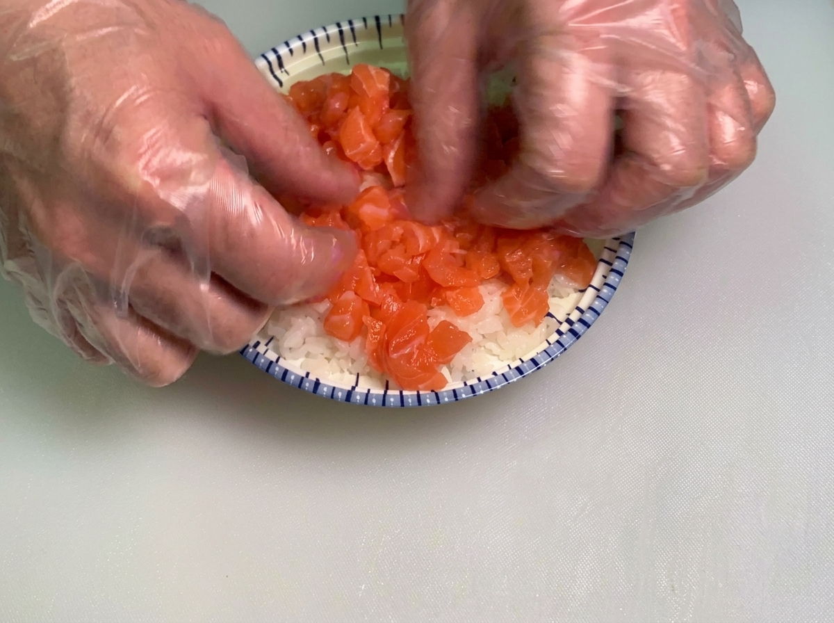 Gloved hands gently spreading a generous layer of diced raw salmon cubes over a bed of white sushi rice in a blue and white patterned bowl.