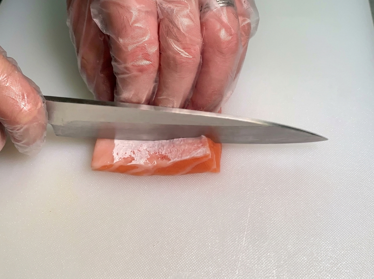 A chef using a knife to carefully slice a small piece of fatty raw salmon belly into thin strips on a white cutting board.