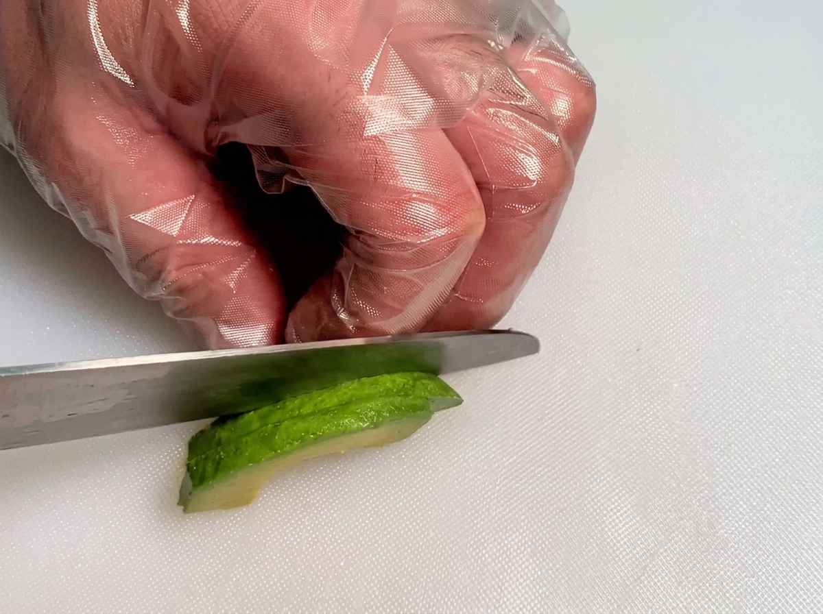 Gloved hands using a knife to dice a slice of fresh green avocado into small cubes.