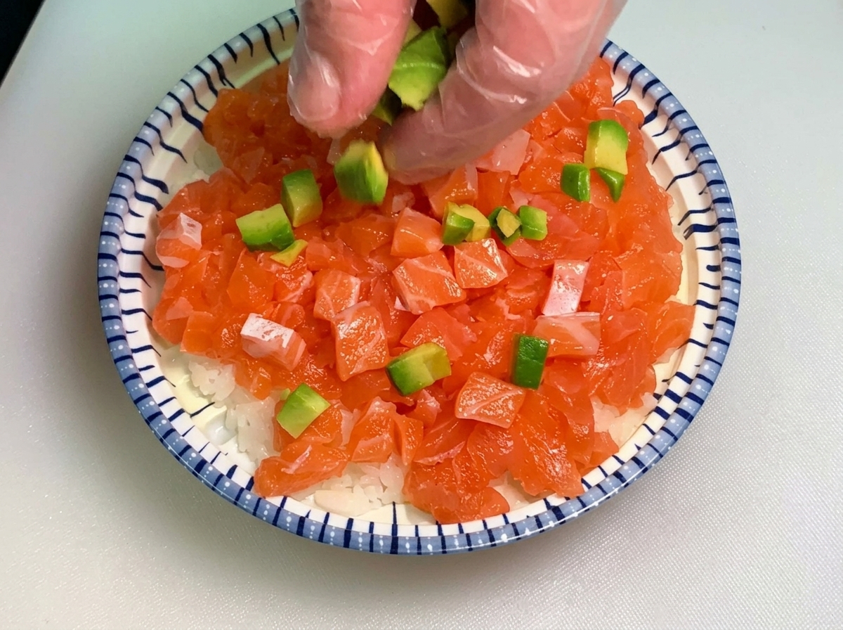 A hand wearing a clear glove sprinkling small cubes of green avocado over a bowl layered with diced salmon and white rice.