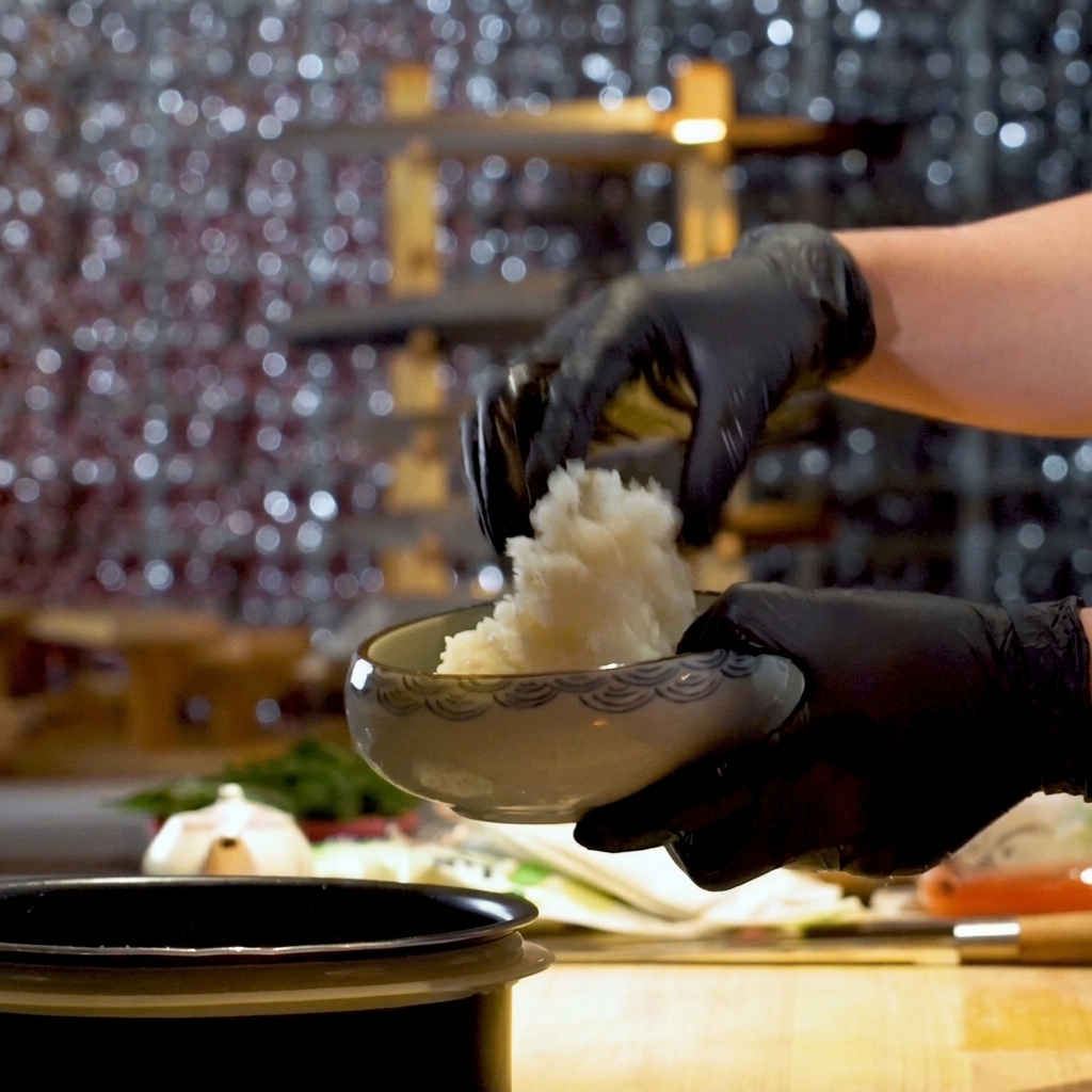 A person wearing black kitchen gloves scooping fluffy white sushi rice into a wave-patterned ceramic bowl.