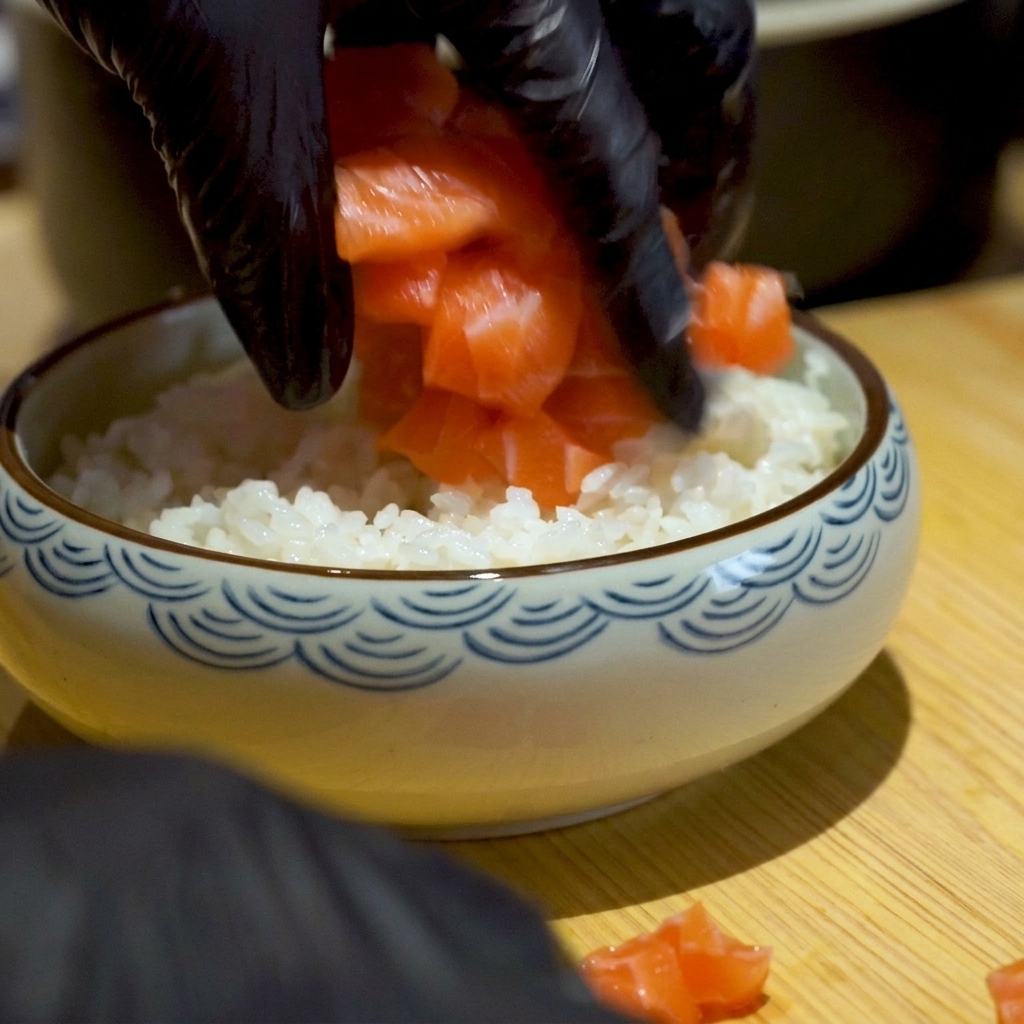 Black-gloved hands placing fresh, vibrant orange diced salmon cubes onto a bed of white rice in a ceramic bowl.