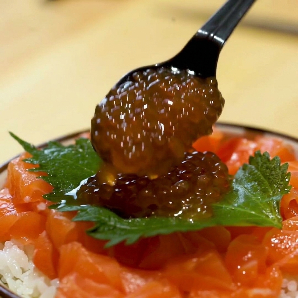 A close-up of a black spoon depositing a heap of glistening orange salmon roe onto a green shiso leaf over a salmon bowl.