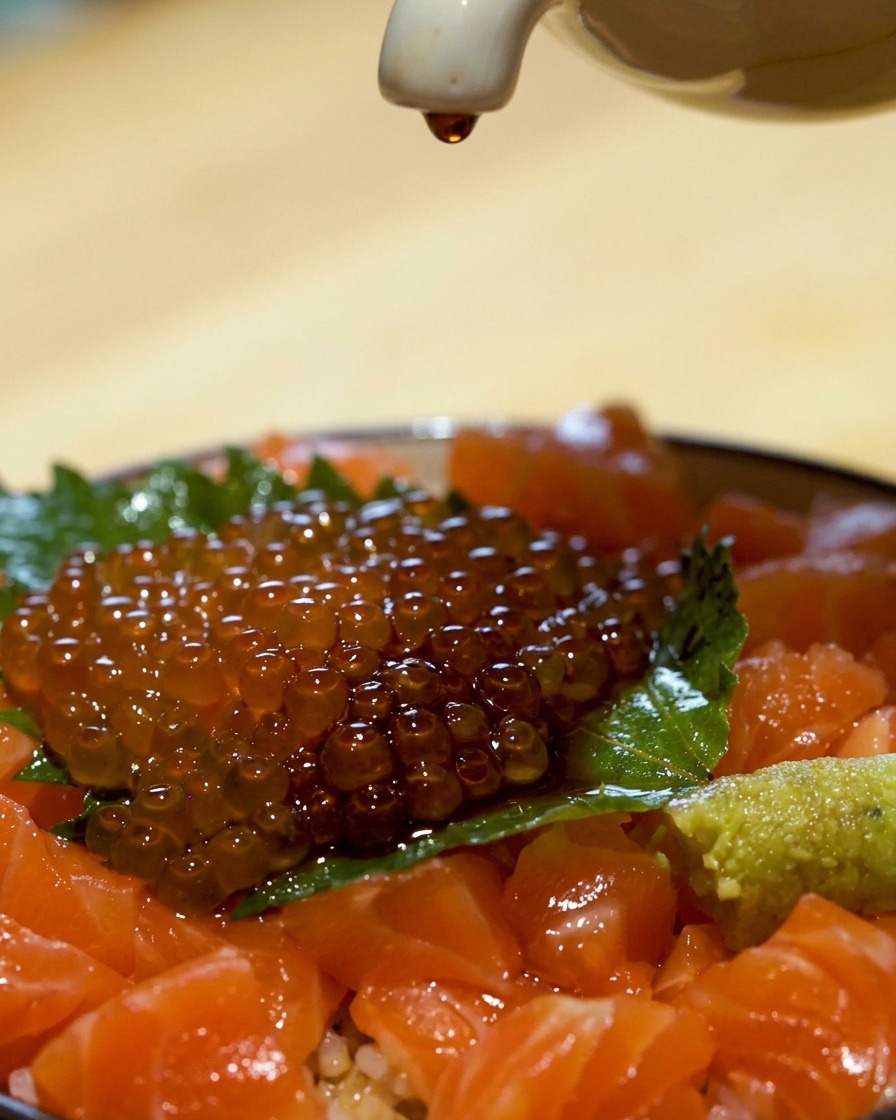 A tight close-up shot of a raw salmon rice bowl featuring fresh cubed salmon, a green shiso leaf, and a heap of glistening orange salmon roe, with a white ceramic spout pouring a drop of dark soy sauce over the top.