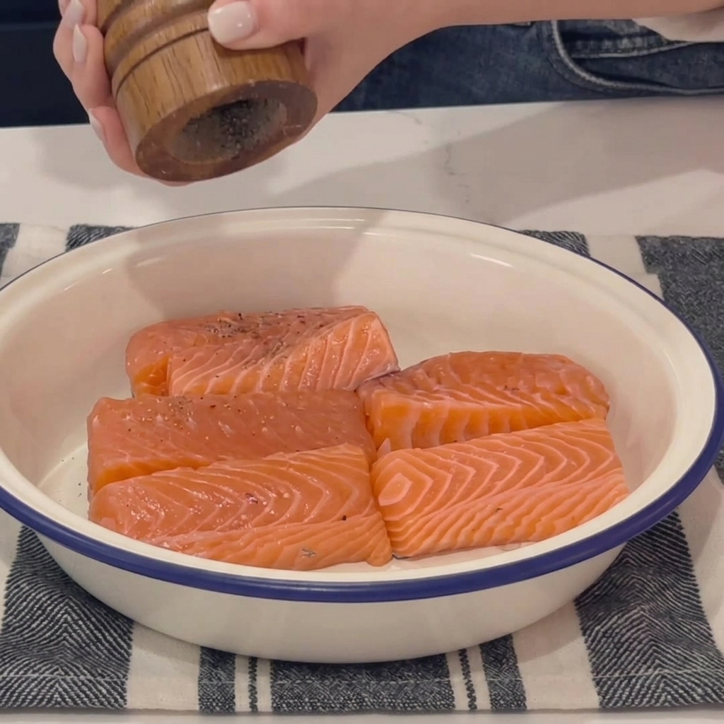 A person uses a wooden pepper grinder to season fresh raw salmon fillets inside a white ceramic bowl.