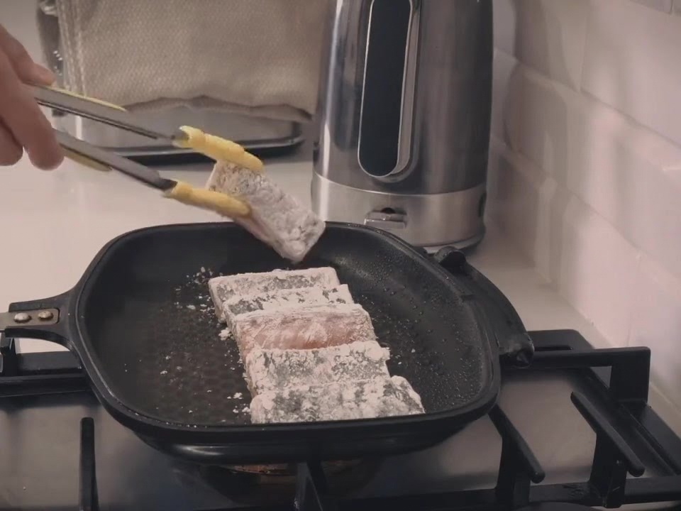 Using yellow-tipped metal tongs to manage and flip multiple salmon fillets frying inside a hot skillet.
