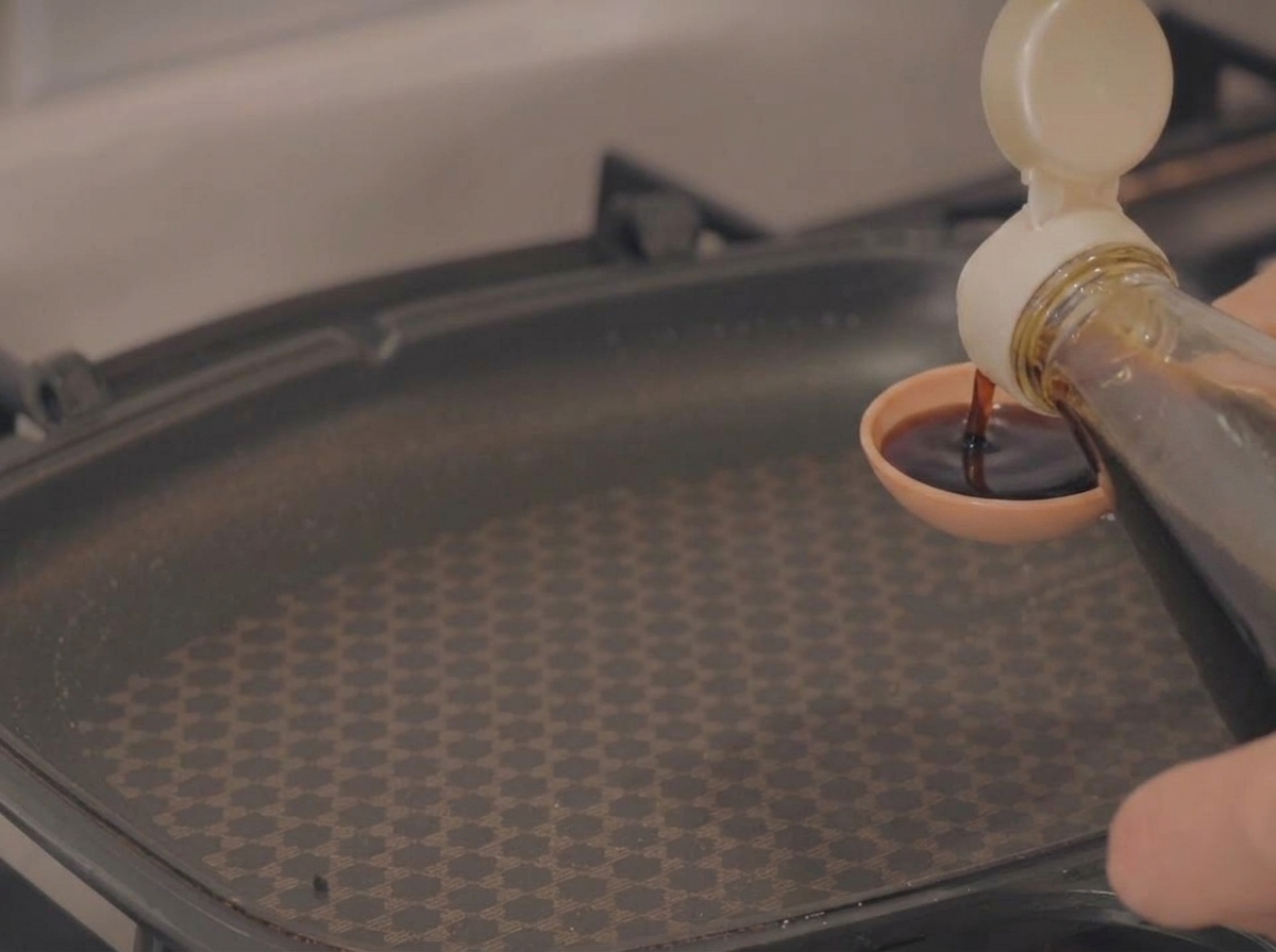 Pouring dark soy sauce from a bottle into a small measuring spoon held above an empty black cooking pan.