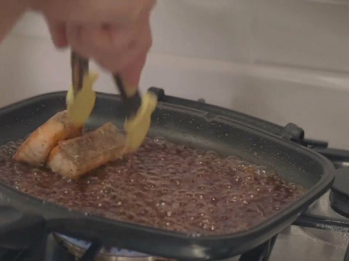 Metal tongs place seared salmon fillets into a square pan filled with a dark, bubbling sauce glaze.