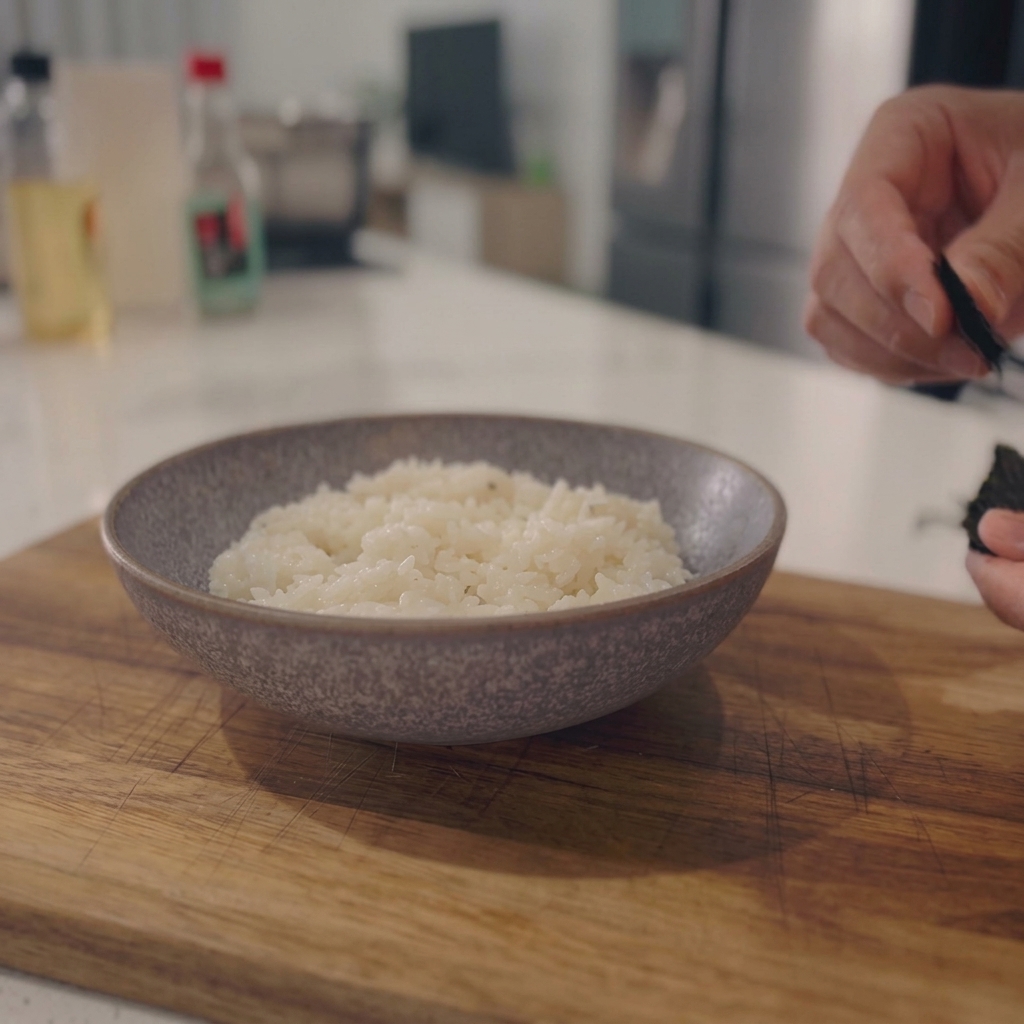 Hands tearing dark sheets of sushi nori seaweed over a ceramic bowl of hot steamed white rice.