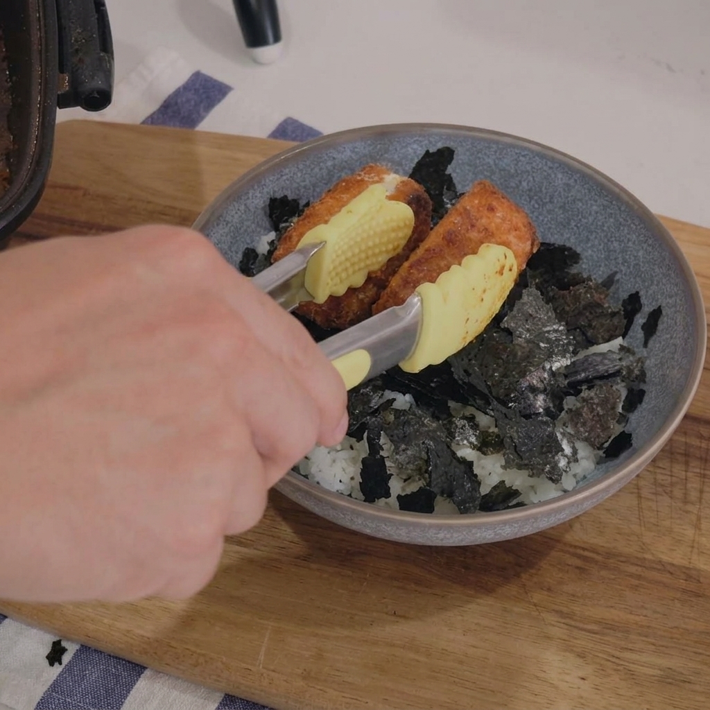 A hand using yellow-tipped tongs to place golden-brown pan-fried salmon fillets into a grey ceramic bowl filled with white rice and shredded nori seaweed.