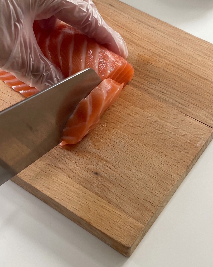 A close-up of a gloved hand using a sharp knife to slice a fresh, orange salmon fillet into thin strips on a light-colored wooden cutting board.