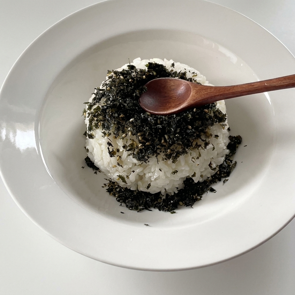 A mound of fluffy white rice in a wide white bowl, topped with a layer of dark green crushed seaweed flakes and a small wooden spoon.