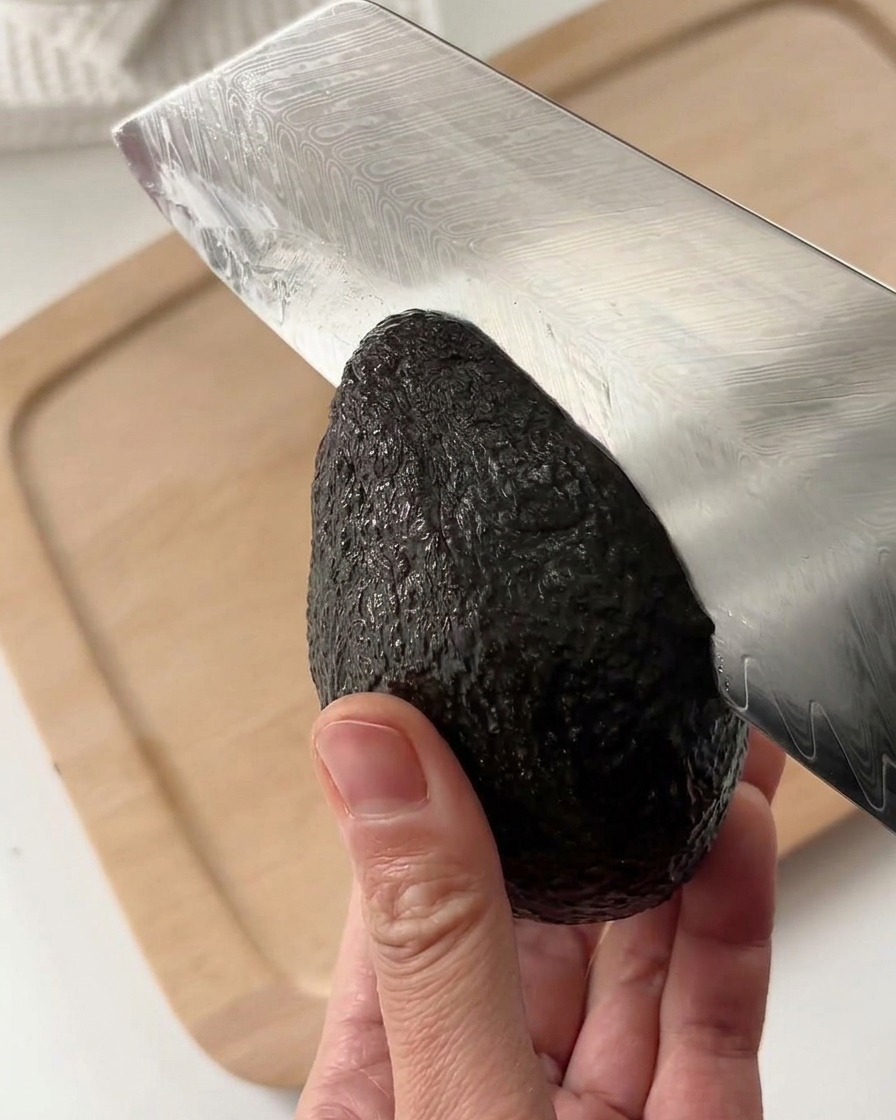 A person cutting a whole, dark green ripe avocado lengthwise with a patterned steel chefs knife on a light wooden background.